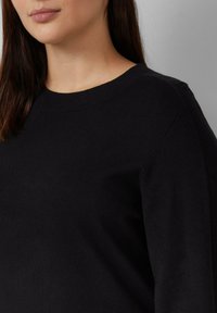 Frau trägt einen schlichten schwarzen langärmligen Pullover mit rundem Halsausschnitt vor einem einfarbigen grauen Hintergrund.