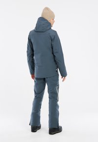 Wasserdichte Skijacke und -hose Set in dunkelblau, mit Kapuze, Reißverschlusstaschen und glatter Textur, kombiniert mit schwarzen Stiefeln.