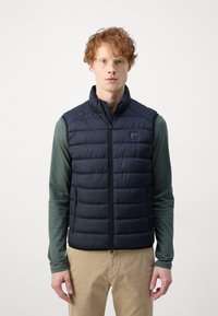 Marineblauer Steppweste mit gequilteter Textur und hohem Kragen. Mit einem Reißverschluss auf der Vorderseite und seitlichen Taschen. Getragen über einem grünen Langarmshirt.