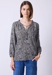Blouse fluide bleu marine avec un motif floral crème, décolletage en V et cordon de serrage. Comprend un col à volants et des manches longues. Associée avec un jean bleu.