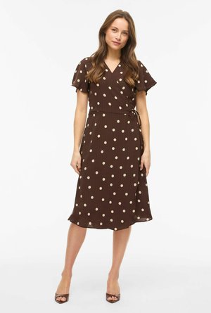 Mujer de pie con un vestido cruzado marrón con lunares beige y mangas acampanadas, combinado con tacones abiertos marrones sobre fondo blanco.