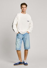 Sudadera blanca de gran tamaño con detalles del logo en negro, combinada con pantalones cortos de mezclilla azul claro. La modelo lleva zapatillas azules con cordones blancos.