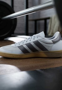 Witte sport schoen met grijze accenten, voorzien van drie horizontale strepen, een gestructureerd oppervlak en een gumrubberen zool. Geplaatst nabij fitnessapparatuur.