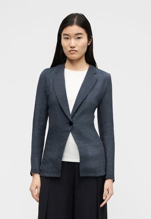 Γυναίκα με μακριά σκούρα μαλλιά, φοράει σκούρο γκρι σακάκι με ένα κουμπί πάνω από λευκή μπλούζα και μαύρο παντελόνι, στέκεται μπροστά σε απλό φόντο.