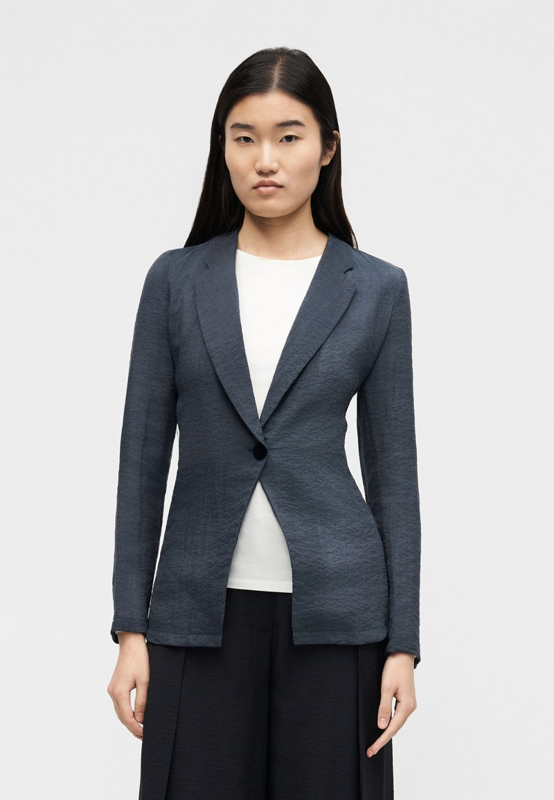 Žena s dugom tamnom kosom, odjevena u tamnosivu jaknu s jednim gumbom preko bijele majice i crnih hlača, stoji nasuprot jednostavnoj pozadini.