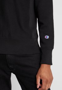 Felpa nera con vita e polsini a costine, caratterizzata da un piccolo logo Champion ricamato sulla manica sinistra sopra una mano rilassata.