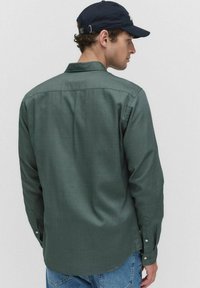 Man in een donkergroen shirt met lange mouwen, blauwe jeans en een zwarte pet, zijwaarts kijkend tegen een neutrale achtergrond.