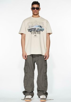 Beige grafisk t-shirt med bildesign, parret med olivenfarvede cargo bukser med flere lommer, og hvide sneakers med orange accenter.
