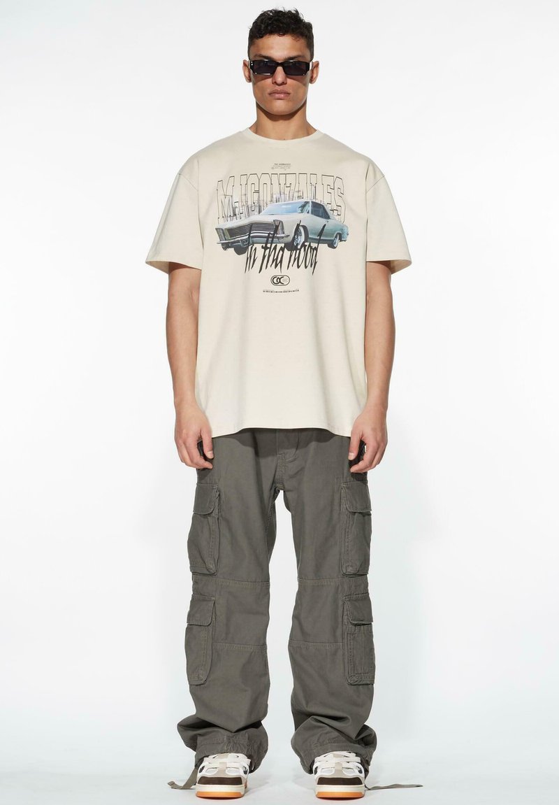 Beige grafisk t-shirt med bildesign, tillsammans med olivgröna cargobyxor med flera fickor, och vita sneakers med orangea detaljer.