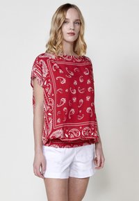 Top in tessuto leggero a fantasia di bandana rossa con maniche corte, caratterizzato da disegni paisley bianchi. Vestibilità ampia con orlo inferiore arricciato.