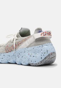 Světle šedá pletená teniska s barevnou modrou stélkou se skvrnami a bílými detaily, s texturovaným povrchem a zadním poutkem na zavěšení.