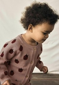 Rosa gestrickte Strickjacke mit roten Erdbeermustern und Holzknöpfen, mit gerippten Bündchen. Das Kind hat lockige Haare und schaut nach unten.