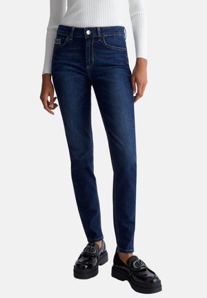 Jeans in denim blu scuro con vestibilità slim, caratterizzati da cuciture a contrasto, chiusura con bottone frontale e tasche posteriori. Abbinati a mocassini neri.