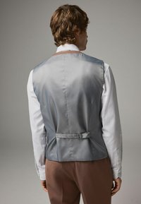 Gilet en satin gris avec une texture lisse, dotée d'une boucle et d'une sangle ajustable à l'arrière, associé à une chemise blanche à manches longues.