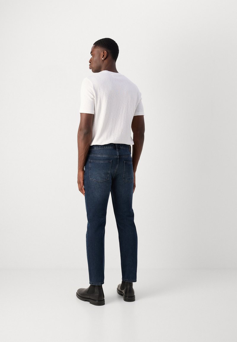 Ciężkie niebieskie dżinsy, dopasowane, średni stan z wyszytymi tylnymi kieszeniami, noszone z białą koszulką z krótkim rękawem i czarnymi botkami za kostkę.