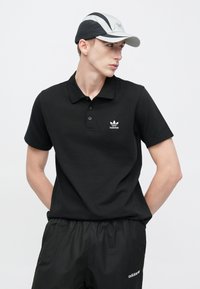 Černé polokošile s třemi knoflíky a bílým logem Adidas. Doplněno lehkými černými šortkami s krátkým střihem, neutrální pozadí.