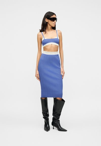 Femme portant un crop top bleu et une jupe midi bordée de blanc, des bottes noires à talons et hauteur genou, et de grandes lunettes de soleil noires, debout devant un fond blanc.