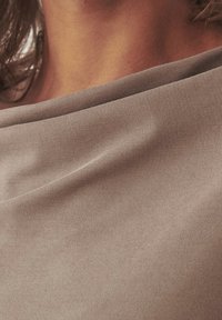 Un tessuto marrone chiaro si drappeggia morbidamente con una superficie liscia, caratterizzato da un design del collo semplice ed elegante con una leggera lucentezza.