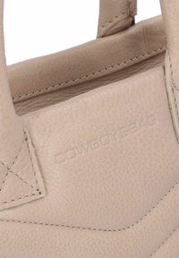 Beige leren handtas met zichtbare stiksels, voorzien van een gestructureerd oppervlak en een debosseerd logo. Het ontwerp omvat twee korte handvatten.