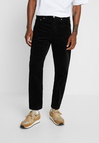 Pantalon en velours côtelé noir avec des côtes verticales, doté d'une braguette à boutons et de poches avant. Porté avec des baskets beiges.