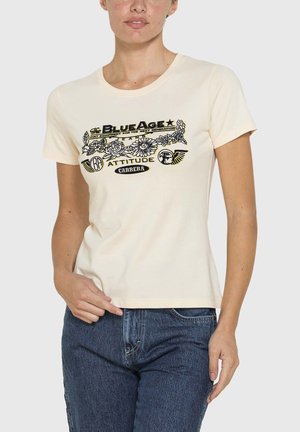 Donna che indossa una t-shirt color crema con motivo floreale nero e giallo e testo, abbinata a jeans di denim blu, in posa con una mano sui fianchi.