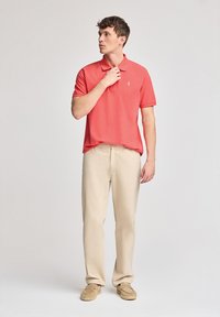Camisa polo roja con cuello y pequeño emblema, combinada con pantalones beige y mocasines marrón claro. El modelo está de pie frente a un fondo liso.