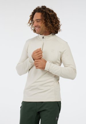 Mand med krøllet hår, der smiler, iført en beige langærmet zip-up trøje og mørkegrønne bukser, mens han justerer sin ærme mod en ensfarvet baggrund.