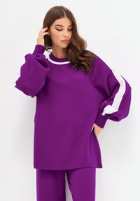 Sweatshirt oversized violet avec des détails en stripe blanche sur les manches et des poignets côtelés. Fabriqué en tissu doux et texturé, il présente un col rond.