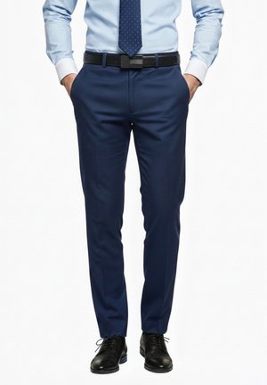 Man wearing blue dress pants, light blue shirt, navy tie with white dots, black belt, and black dress shoes, hands in pockets.