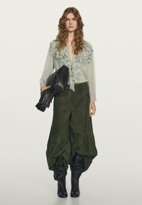 Jasnozielona bluzka z falbanami i głębokim dekoltem w kształcie litery V, zestawiona z ciemnozielonymi, szerokimi spodniami z zamszu oraz czarnymi, sięgającymi kolan butami, trzymając czarną kopertówkę.