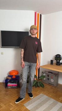 Pruun graafiline T-särk lühikeste varrukatega, heledast hallist sirge lõikega teksad, mustad libisemisjalanõud, seistes kaunistatud ruumis, kus on taimi.