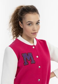 Fleece varsityjackets in felroze met witte mouwen. Voorzien van een grote paarse letter "M" en zilveren drukknoopjes aan de voorkant.