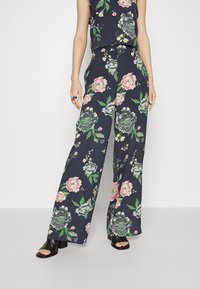 Calças largas florais em azul-marinho com flores rosas, verdes e brancas; tecido leve; combinadas com saltos abertos pretos e uma blusa correspondente.