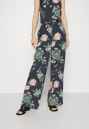 Calças largas florais em azul-marinho com flores rosas, verdes e brancas; tecido leve; combinadas com saltos abertos pretos e uma blusa correspondente.