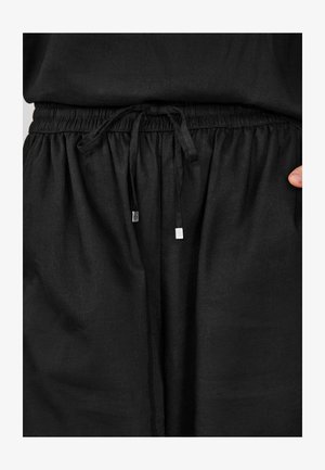 Crne hlače od lana s elastičnim pojasom i vezicom, vrhovi vezice obloženi srebrnim završecima, te krojem širokog kroja. Glatka tekstura s mogućnošću laganog pada tkanine.