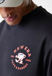 Black cotton t-shirt with a round red and white graphic featuring a cup character and the text "NEW ERA CAFE & BAKERY" in bold letters.