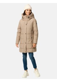 Beige puffer coat met een capuchon, voorzien van een gequilt ontwerp, knopen aan de voorkant, ritszakken aan de zijkant, gecombineerd met blauwe jeans en witte sneakers.