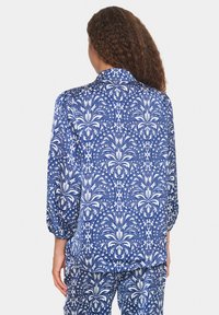 Blusa blu con motivo floreale bianco, maniche lunghe, colletto rotondo e texture morbida. Presenta una vestibilità ampia e leggeri arricciamenti alle maniche.