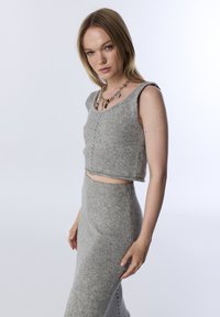 Kobieta ubrana w bezrękawnikowy szary sweter w cropie oraz dopasowaną spódnicę o wysokim stanie, ozdobiona grubym naszyjnikiem z łańcuchem i zawieszkami.