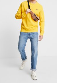 Sweat-shirt jaune à col ras du cou, jean bleu et baskets blanches. Un sac bandoulière marron avec une fermeture éclair repose sur l'épaule. Tenue décontractée.