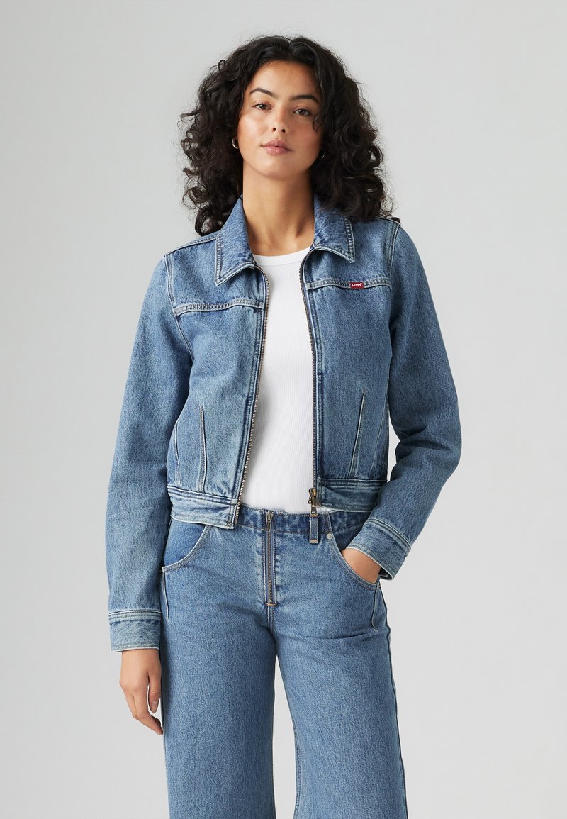 Veste en denim bleu avec fermeture éclair à l'avant, col pointu et deux poches latérales. Présente une petite étiquette de marque rouge sur la poitrine. Texture lisse.