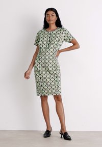 Grünes gemustertes Kleid mit kurzen Ärmeln, das ein geometrisches Design, schwarze Abschlüsse und eine knielange Silhouette aufweist. Getragen mit schwarzen Schuhen.