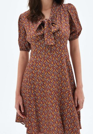 Femme portant une robe à manches courtes de couleur rouille avec un motif cachemire orange et blanc et un nœud à l'avant au niveau de l'encolure.