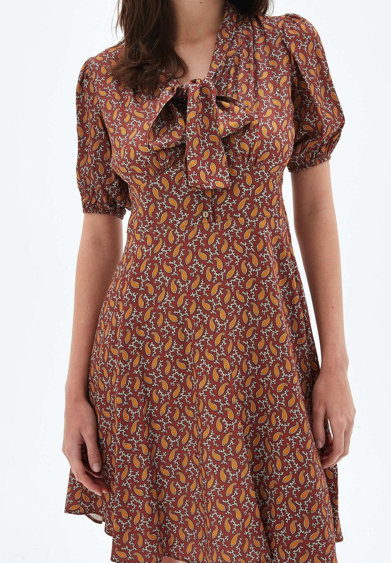 Femme portant une robe à manches courtes de couleur rouille avec un motif cachemire orange et blanc et un nœud à l'avant au niveau de l'encolure.