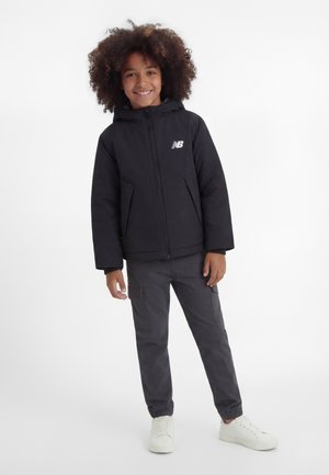 Smilende barn med krøllet hår iført sort hættetrøje, mørke cargobukser og hvide sneakers, stående mod en hvid baggrund.