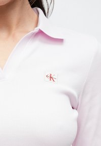 Polo rosa chiaro a costine con colletto, caratterizzata da una toppa con il logo Calvin Klein in rosso e bianco sul petto. Tessuto liscio, design aderente.
