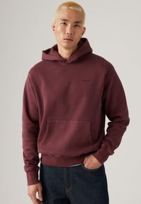 Maroon hoodie made of cotton with a front pocket, ribbed cuffs, and hem. Features a subtle logo on the chest, worn with dark jeans.