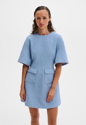 Kvinde med vådt hår iført en lyseblå, kortærmet kjole med tekstur og to lukkelommer foran, stående foran en ensfarvet hvid baggrund.