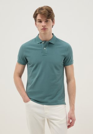 Junger Mann, der ein türkisfarbenes kurzärmeliges Poloshirt und weiße Hose trägt, steht mit einer Hand in der Tasche vor einem einfachen Hintergrund.