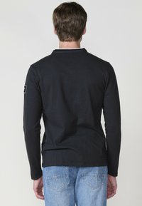 Hosszú ujjú fekete ing, struktúrált anyagból, kerek nyakkivágással és diszkrét folttal a bal ujján, világoskék farmerral párosítva.
